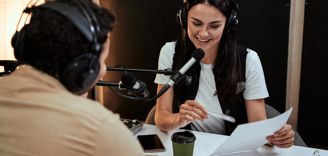 Portait of happy female radio host smiling, reading a script from paper while talking to male guest, presenter, moderating a live show in studio