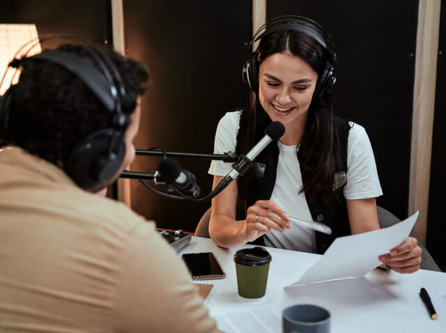 Portait of happy female radio host smiling, reading a script from paper while talking to male guest, presenter, moderating a live show in studio